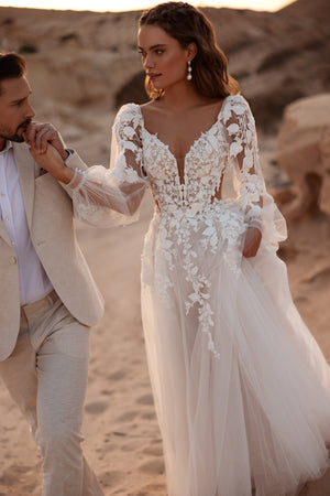 Das Alia-Brautkleid von DAMA Couture hat ein figurbetontes Spitzenkorsett mit 3D-Blütenmotiven und ein tiefes V-Ausschnitt, das die Taille zart formt.