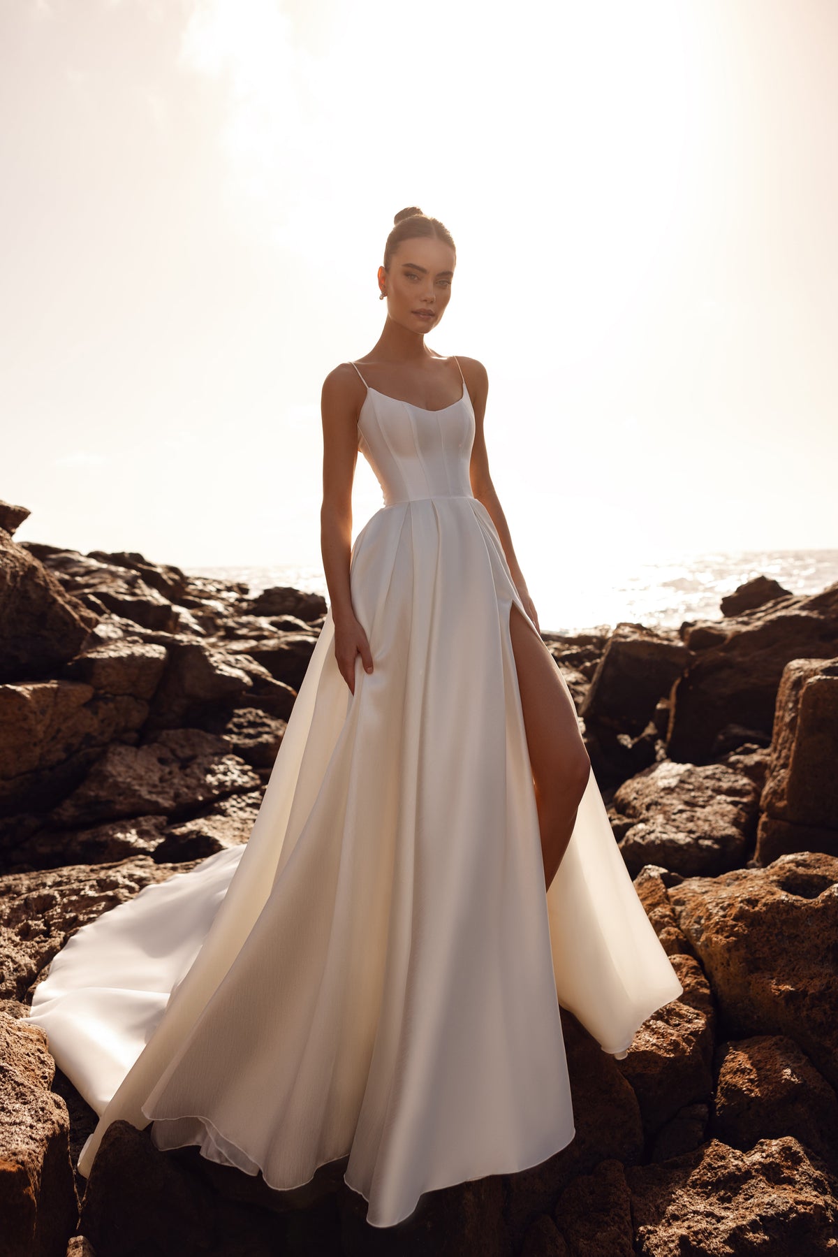 Brautkleid Alya aus mattem Satin mit schmalen Trägern und hohem Beinschlitz, Vorderansicht am Meer