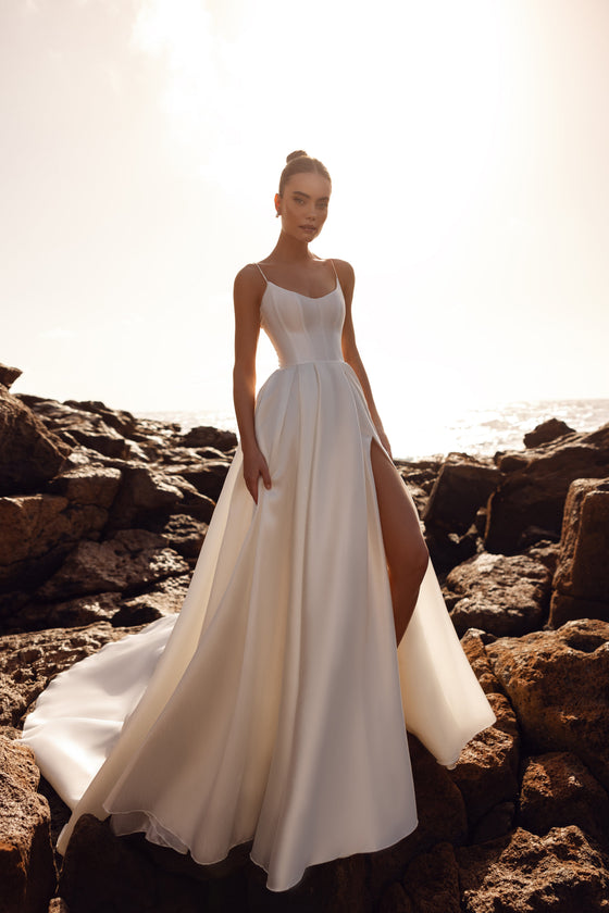Brautkleid Alya aus mattem Satin mit schmalen Trägern und hohem Beinschlitz, Vorderansicht am Meer