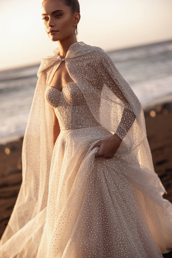 Brautkleid Amira in A-Linie aus Glitzertüll mit offenem Kapuzencape, Dreiviertelansicht am Strand