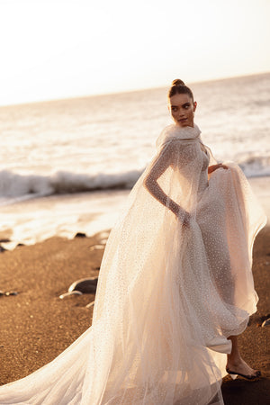 Brautkleid Amira mit wehendem Cape in ganzer Silhouette vor dem Meer