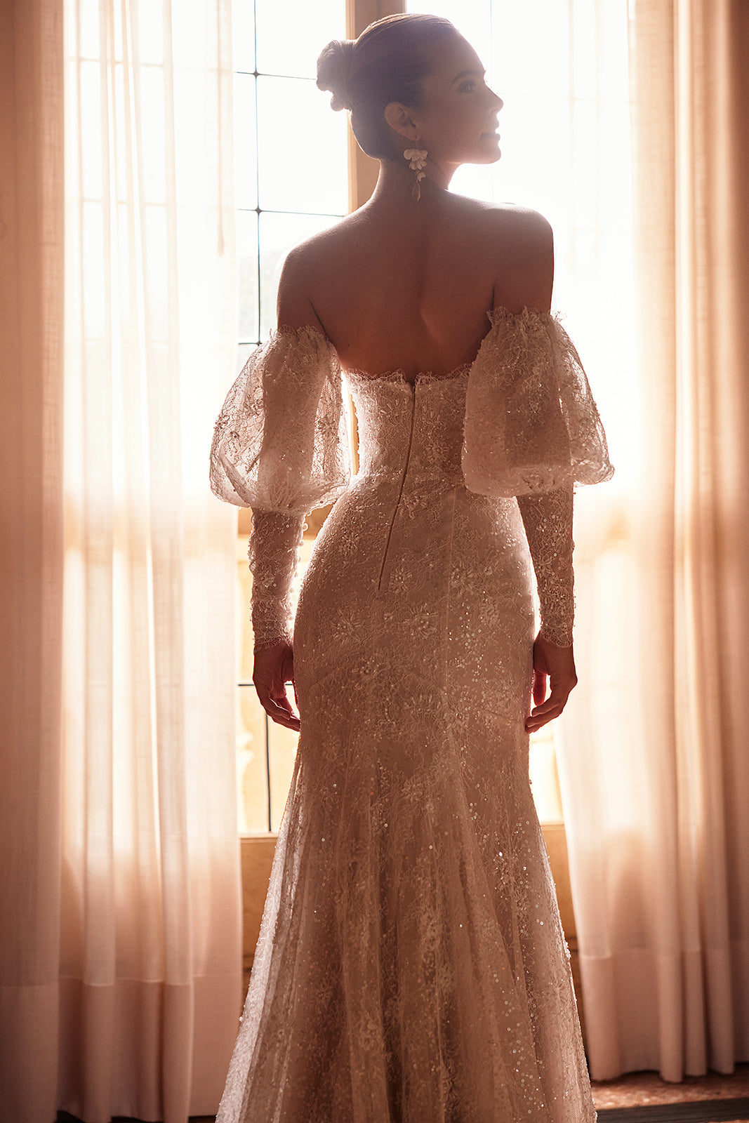 Brautkleid Genevieve im Gegenlicht am Fenster, offener Rücken und abnehmbare Puffärmel