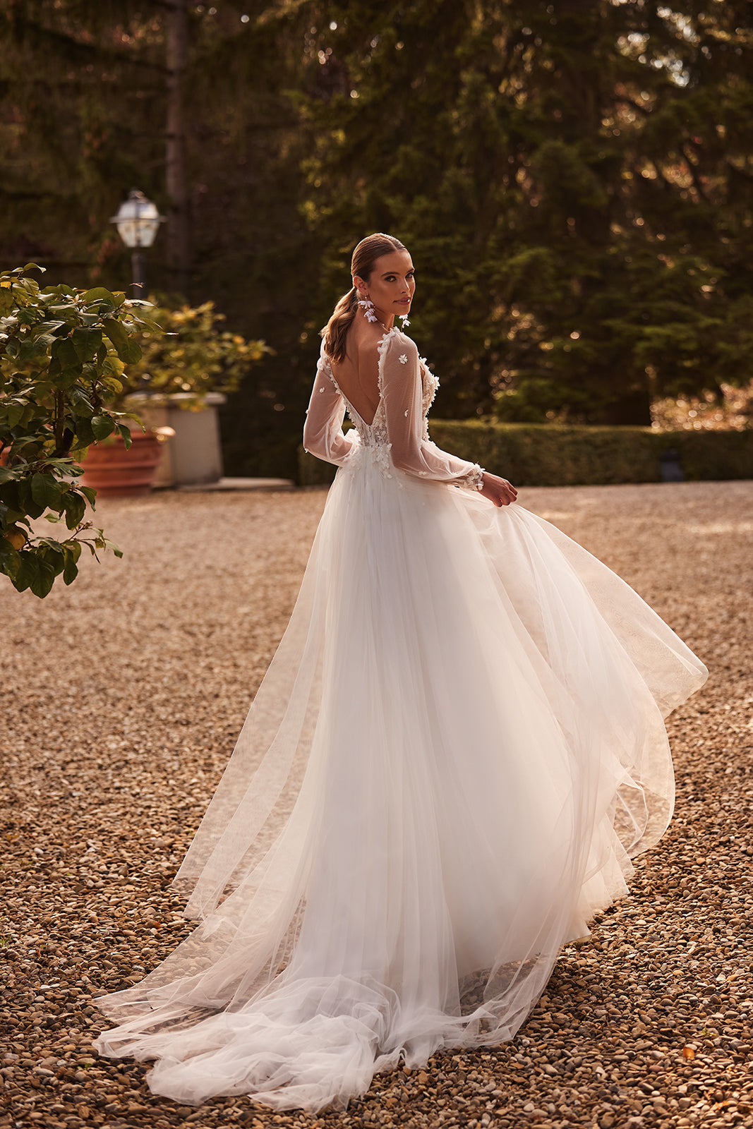 Brautkleid Nozomi von hinten, ganze Silhouette mit Tüllschleppe auf dem Kiesweg