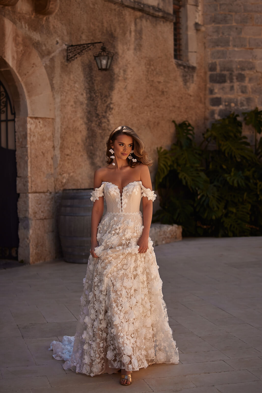 Brautkleid Olivia von vorn im Innenhof, schulterfreier Herzausschnitt und 3D-Blüten am Rock