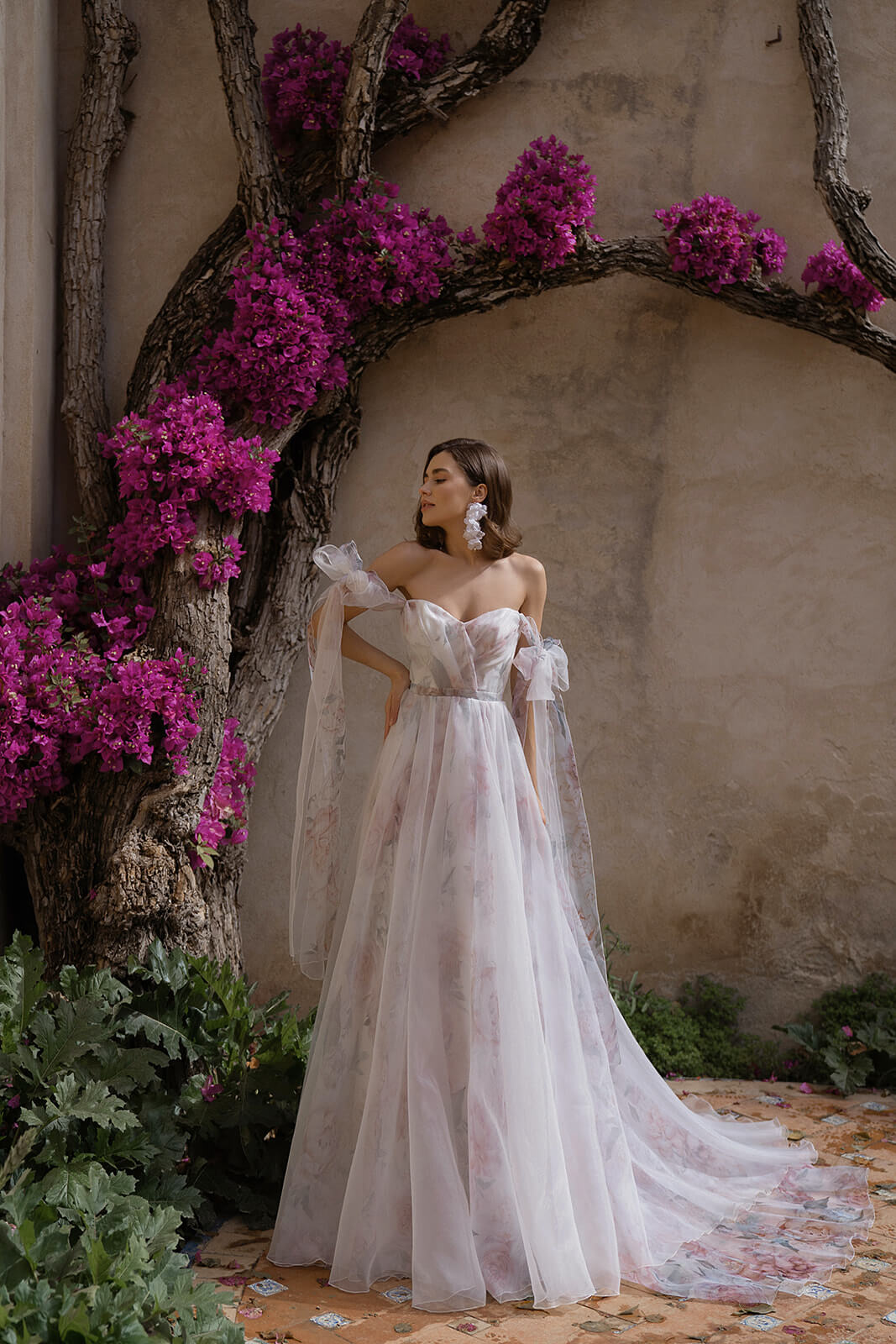 Brautkleid Peony, ganze Silhouette vor der blühenden Mauer mit Schleppe