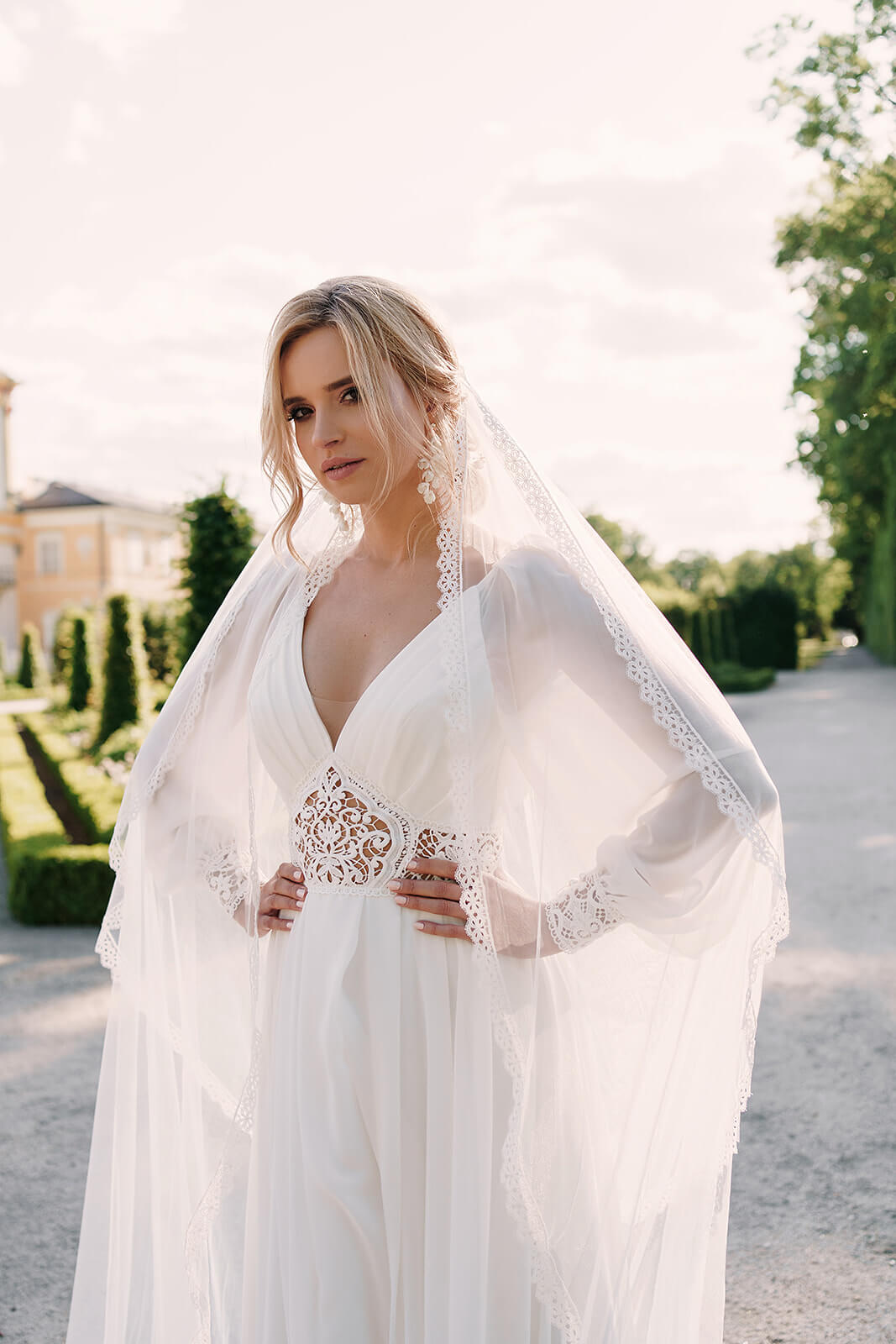 Brautkleid Safona mit Schleier von vorn, Hände in der Taille im Schlosspark