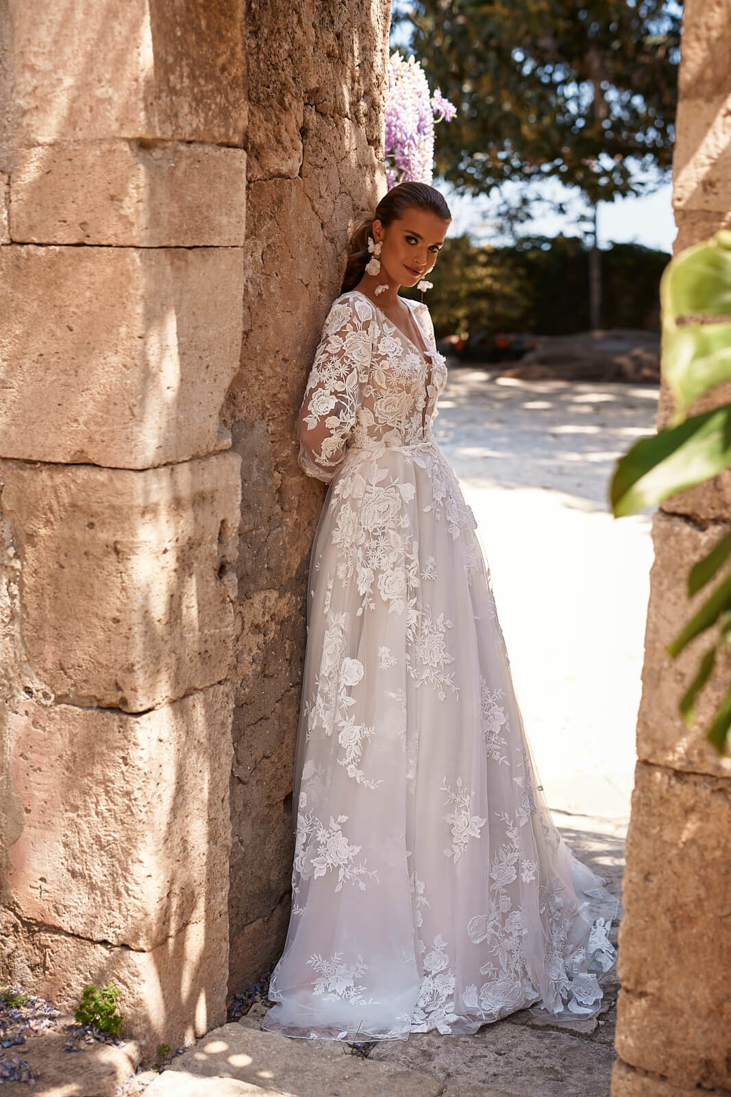 Brautkleid Asami in A-Linie, ganze Silhouette seitlich an der Steinmauer, Spitzenschleppe