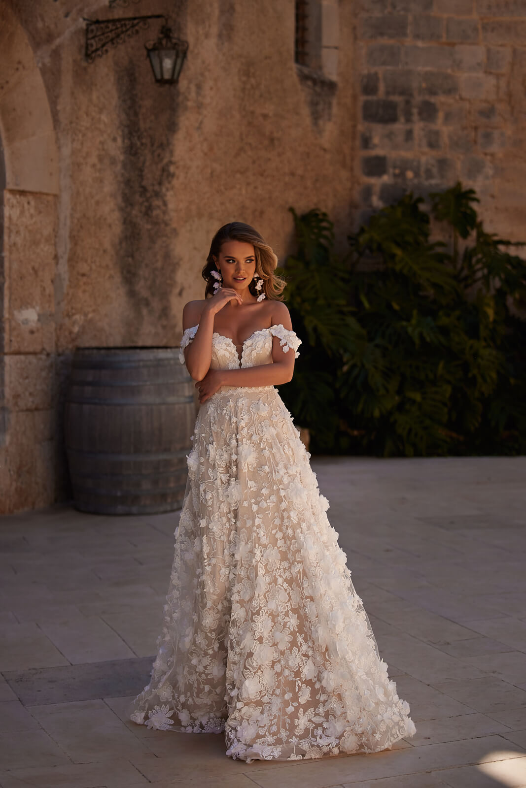 Brautkleid Olivia in ganzer Figur, A-Linie mit 3D-Blüten vor einer Steinmauer
