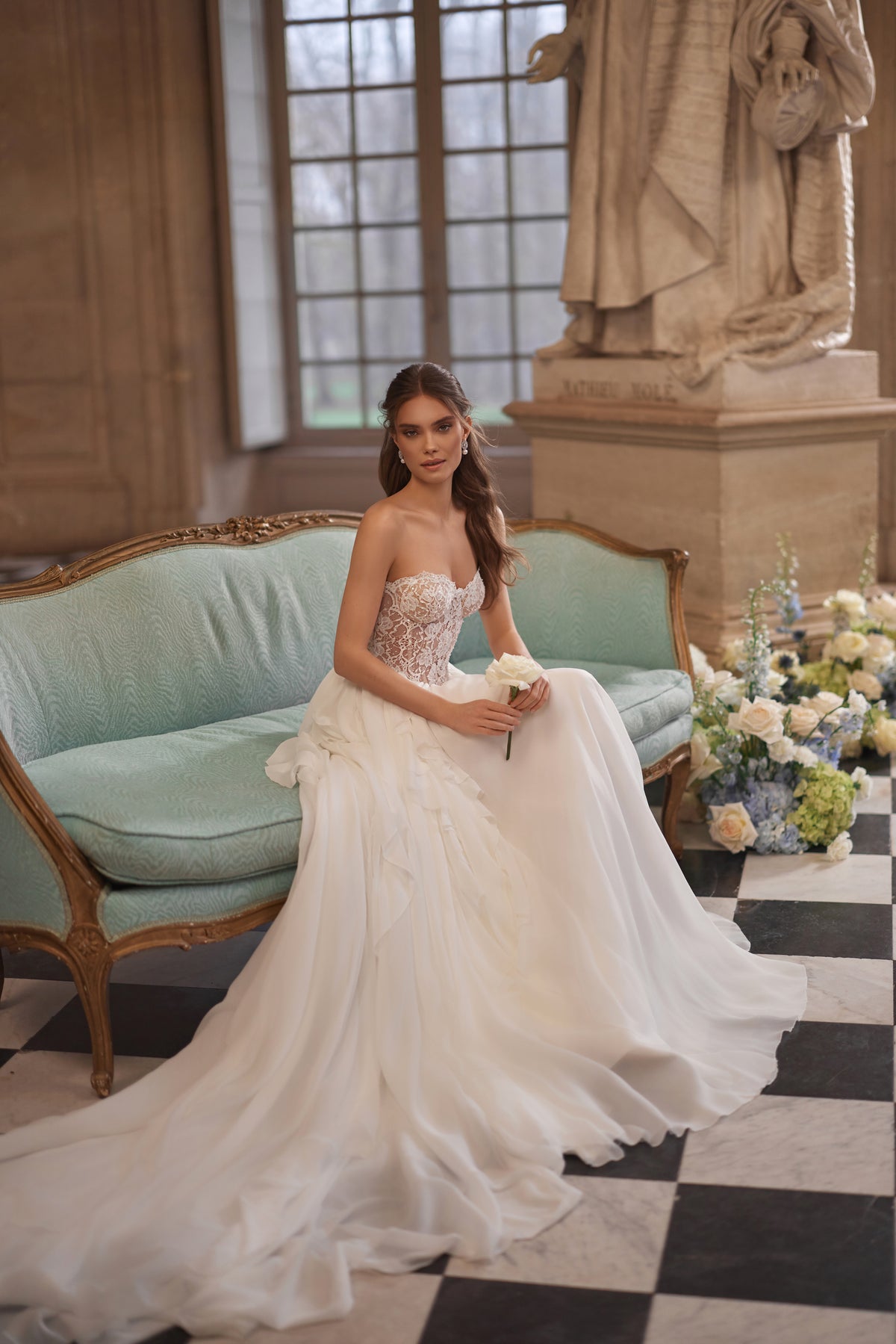 Brautkleid Isolde in A-Linie – trägerlose Spitzencorsage und Volantrock, ganze Figur auf dem Sofa