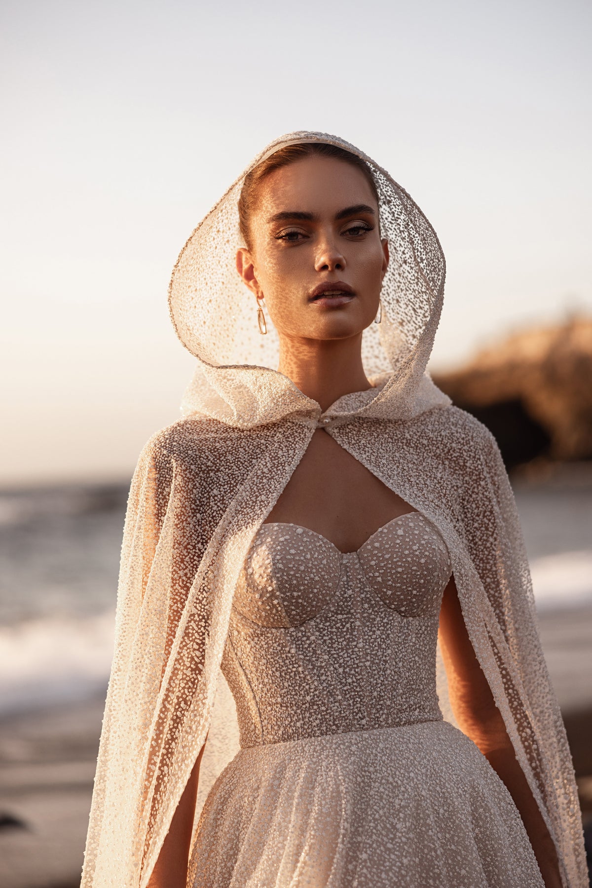 Brautcape Amira mit Kapuze aus glitzerndem Tüll über der Corsage, Halbfigur am Meer