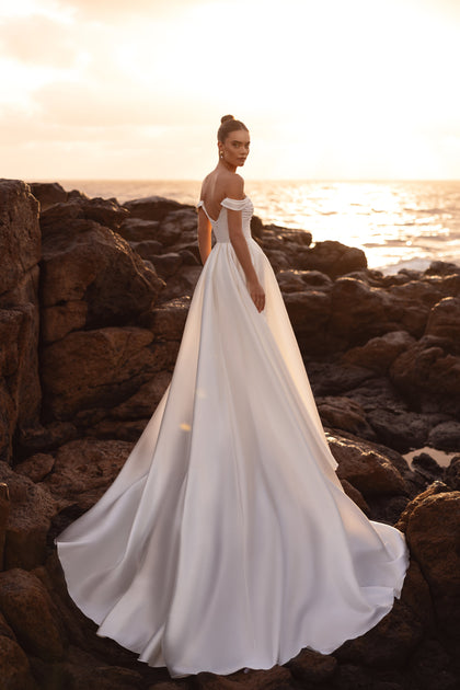 Brautkleid Mellisandre mit schulterfreiem Ausschnitt und weiter Schleppe, die im Sonnenuntergang erstrahlt.