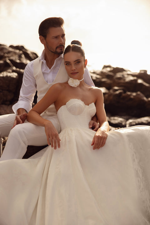 Das Brautkleid Cholly mit Korsettoberteil und weitem Rock, romantisch zwischen Meeresfelsen aufgenommen.