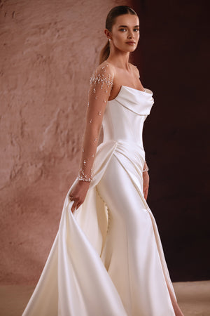 Das Brautkleid Aylin mit transparenten Ärmeln und Perlenbesatz sowie drapiertem Ausschnitt verkörpert zeitlose Eleganz.