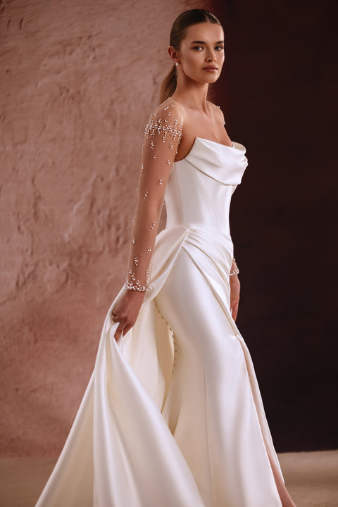 Das Brautkleid Aylin mit transparenten Ärmeln und Perlenbesatz sowie drapiertem Ausschnitt verkörpert zeitlose Eleganz.