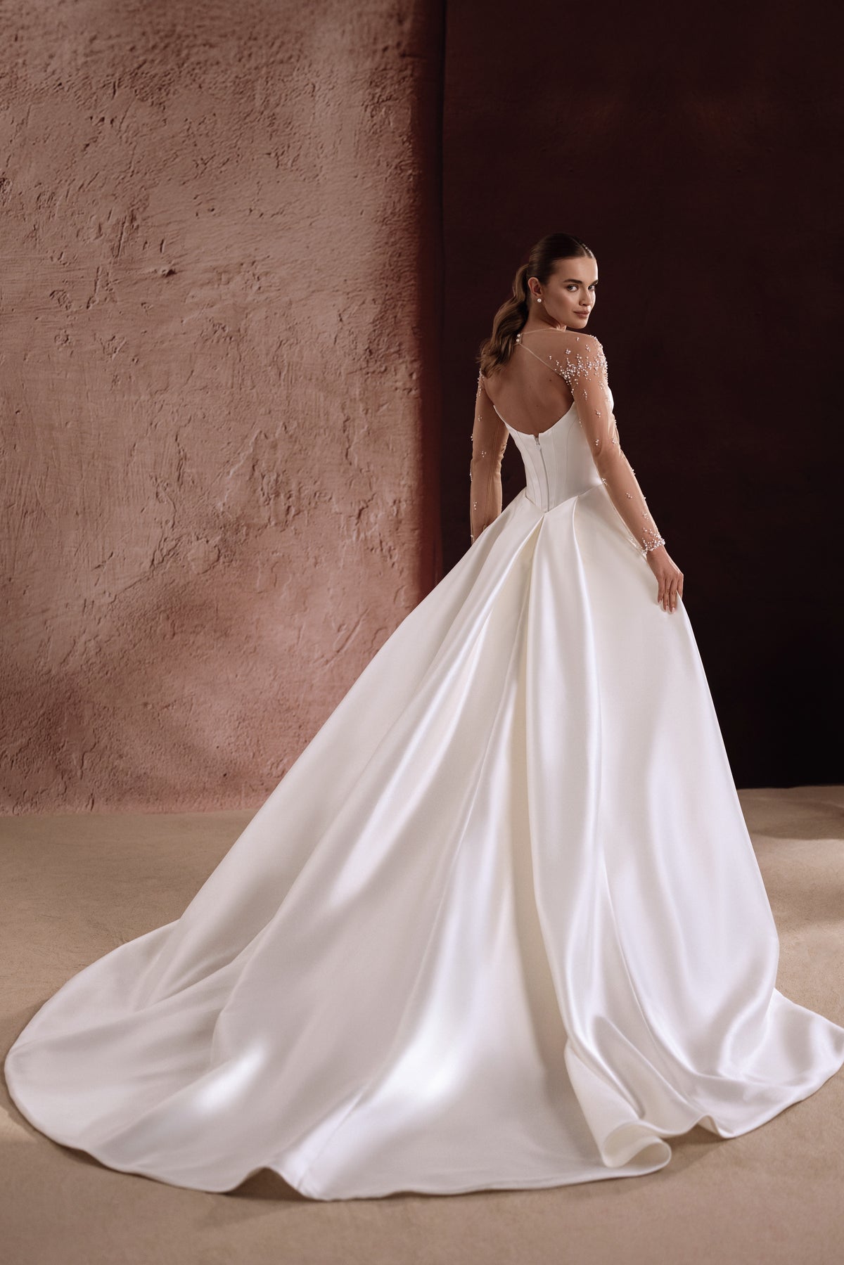 Brautkleid Aylin von hinten, weiter Satin-Überrock mit Schleppe vor einer Lehmwand