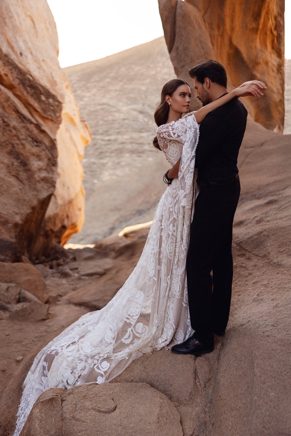 Brautkleid Dune mit langer Spitzenschleppe auf Wüstenfelsen, Braut neben dem Bräutigam
